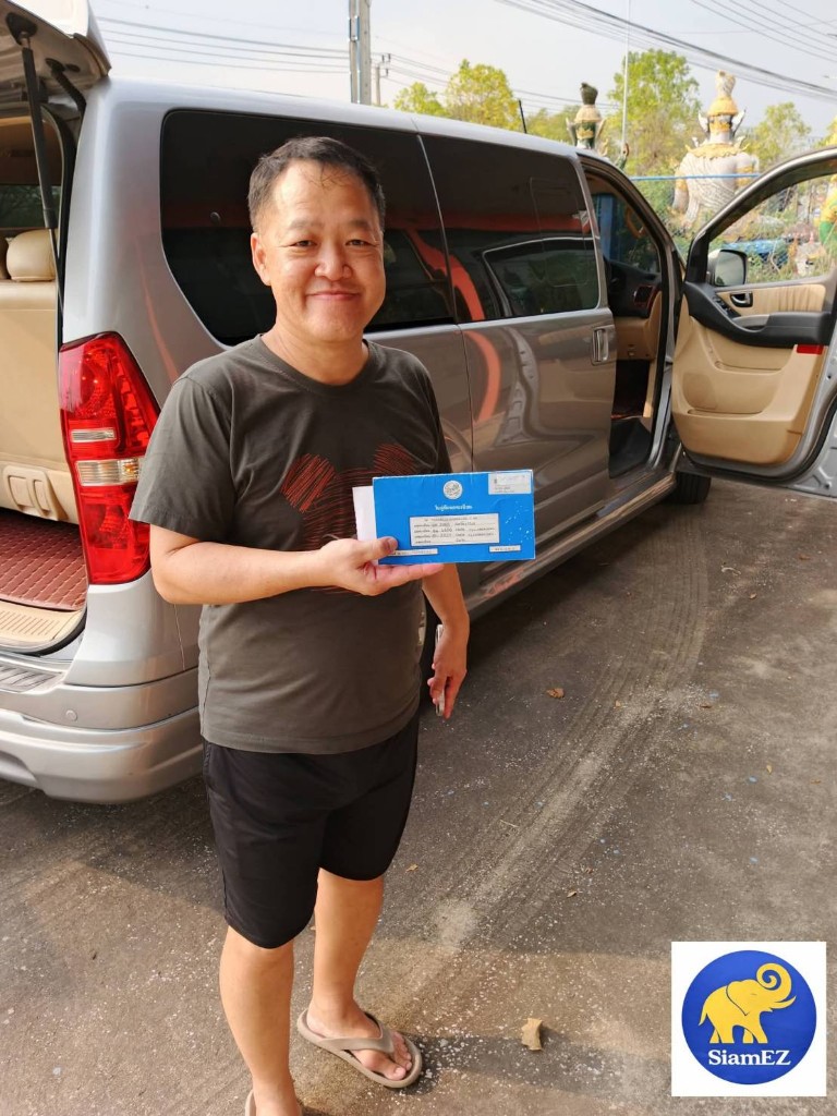 Customer holding blue Thai vehicle registration booklet in front of a silver minivan