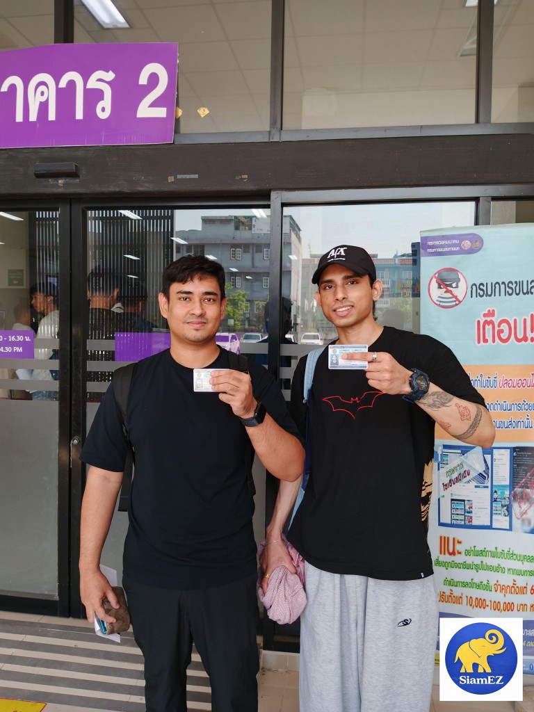 Two men proudly holding their newly obtained Thai driver's licenses in front of a licensing office.