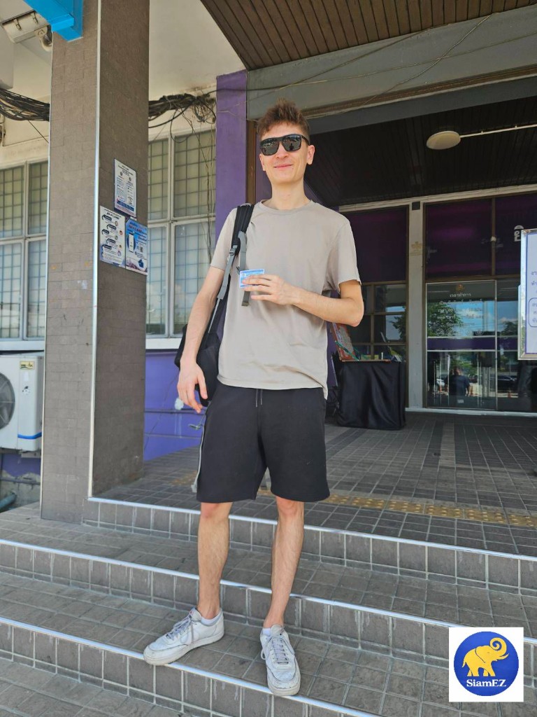 A man smiling and holding his new Thai driver's license in front of a government building.